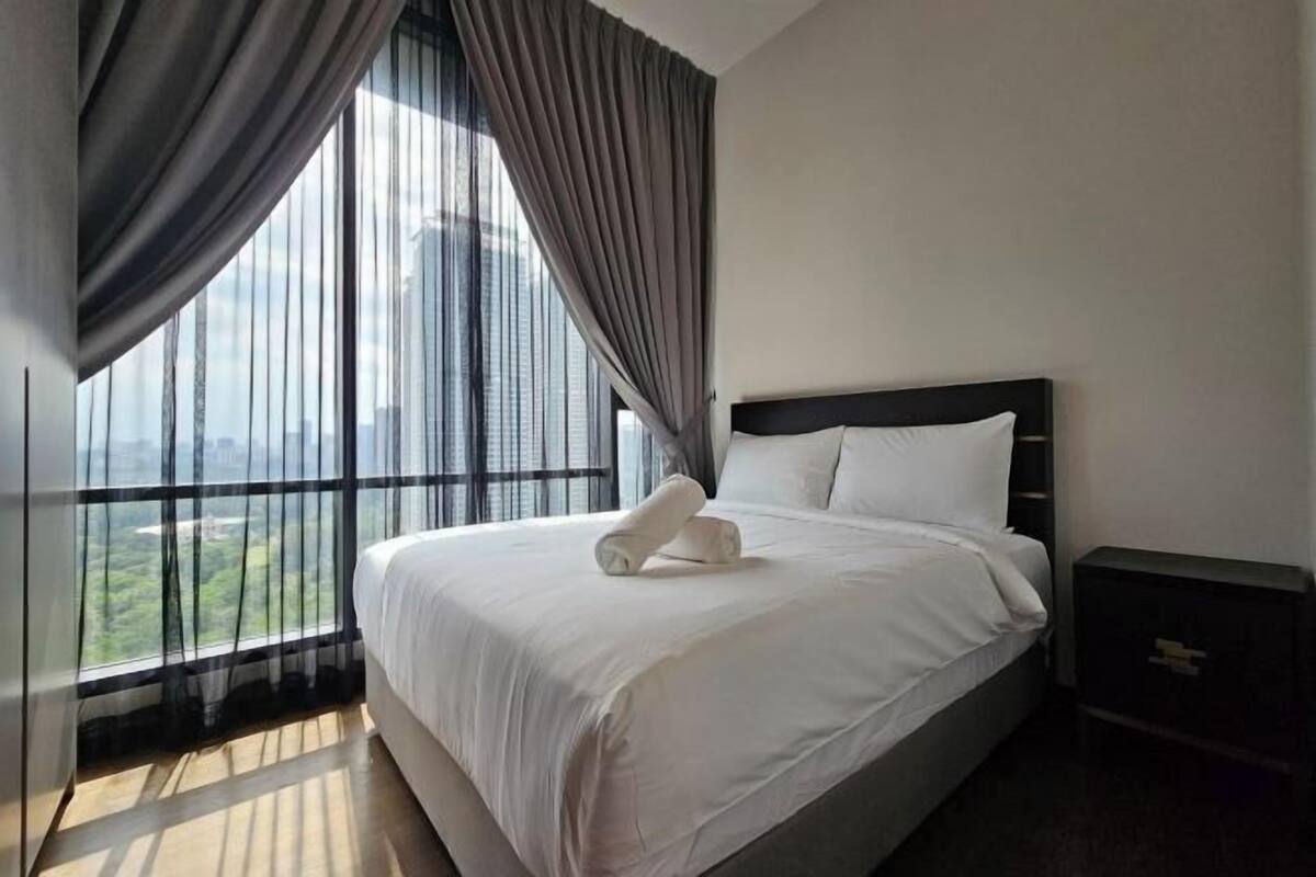 Une chambre d’hôtel avec un grand lit, une table de chevet et une vue sur le paysage urbain, visible à travers de fines rideaux.