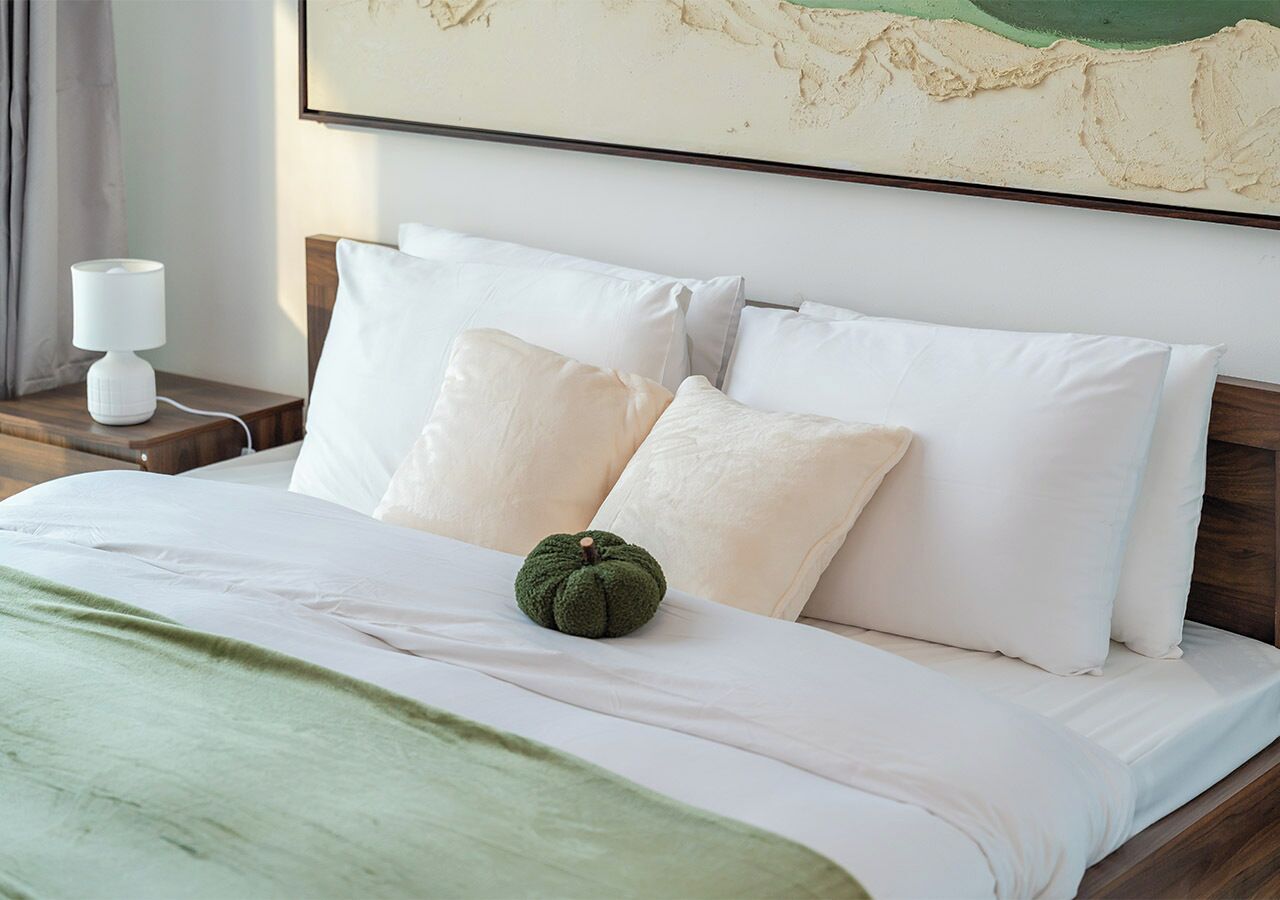 Un lit bien fait, avec des draps blancs, un plaid vert et une citrouille décorative posée sur les oreillers.