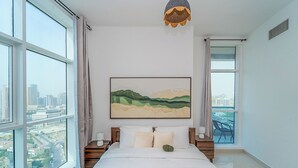 A bedroom with a large bed, a city view, a painting on the wall, and a unique ceiling light. - Lux JVT Resort Pools Spa Gym Kids Play (Dubai)