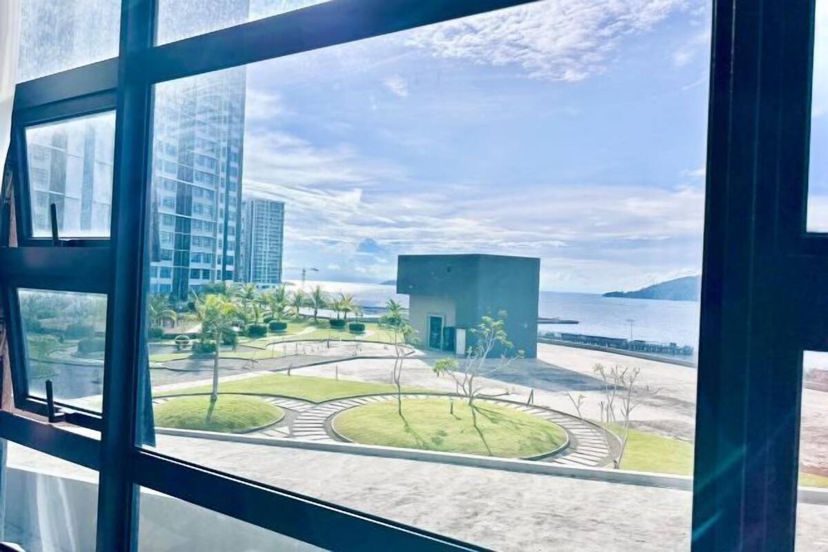 A modern building with large windows, a view of a landscaped area with pathways and trees, and a body of water in the distance.