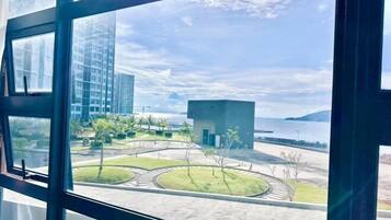 A modern building with large windows, a view of a landscaped area with pathways and trees, and a body of water in the distance.