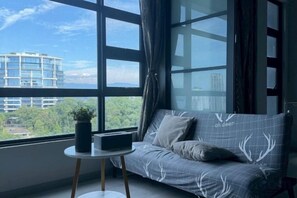 A modern room with a sofa facing a large window, a small table with a plant, and a cityscape view. - Jesslton quay Dream stay (Kota Kinabalu)