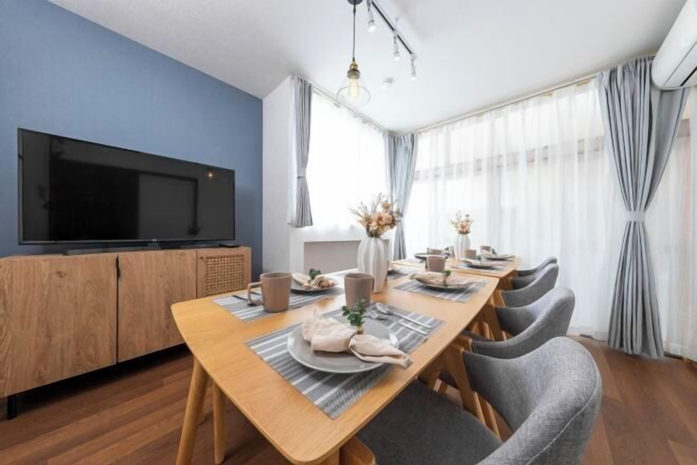 A dining area with a wooden table, grey chairs, a television on a wooden cabinet, and a window with sheer curtains.