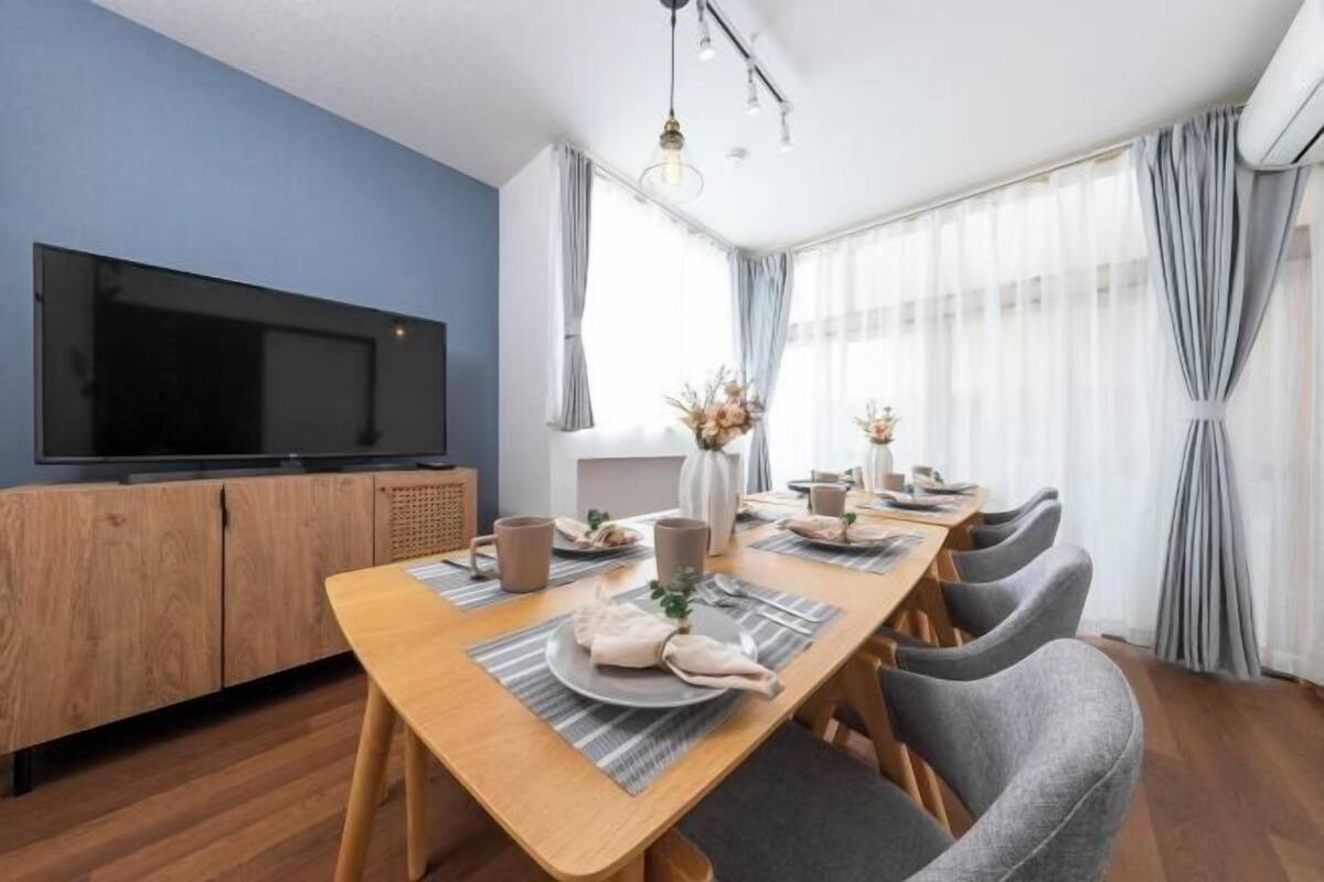 A dining area with a wooden table, grey chairs, a television on a wooden cabinet, and a window with sheer curtains.