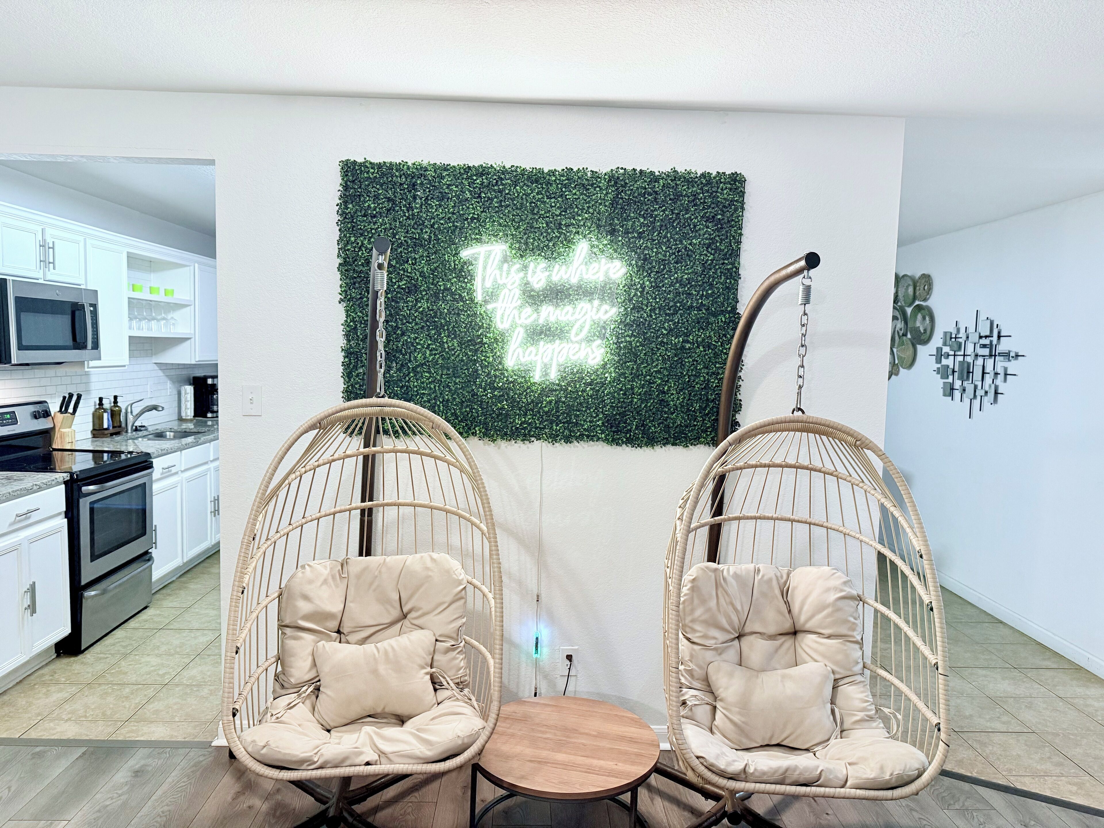 A modern kitchen with white cabinets, a microwave, and a coffee machine. There are two wicker chairs with cushions and a small wooden table. A neon sign on the wall reads "This is the magic happens."