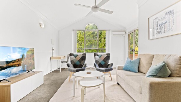 A modern living room with a ceiling fan, a large TV, two armchairs, a sofa, and a window with a view of trees.
