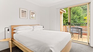 A bedroom with a wooden bed, white bedding, a nightstand, a desk, and a sliding glass door to an outdoor patio with chairs and a table.