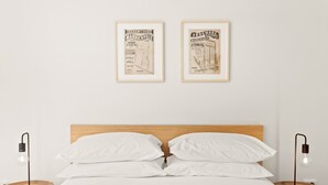A neatly made bed with white linens, two framed historical posters on the wall, and two bedside tables with lamps.