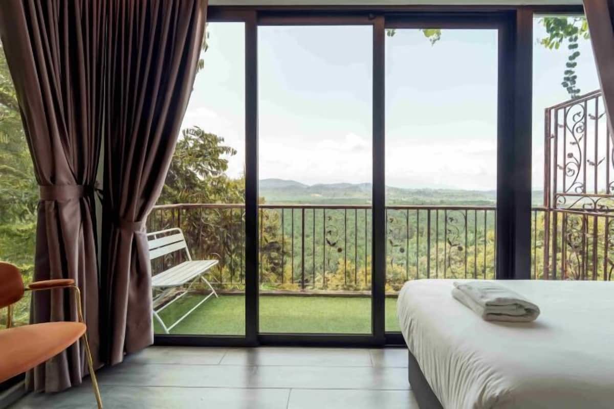 A bedroom with a large sliding glass door leading to a balcony with a view of greenery and hills.