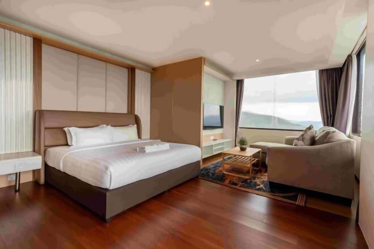 A modern hotel room with a large bed, a sofa, a small table, and a large window offering a view.