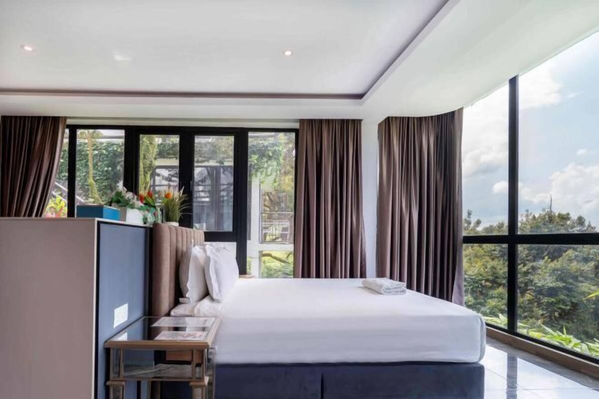 A modern bedroom with a large bed, a bedside table, and a view of greenery through large windows.