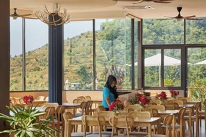 A dining area with tables and chairs, large windows with a view of hills, and a ceiling fan. - Bilut Hills - Eco Resort (Teras)