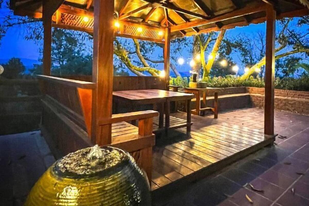 An outdoor wooden gazebo with a table and benches, surrounded by a stone wall and illuminated by string lights.