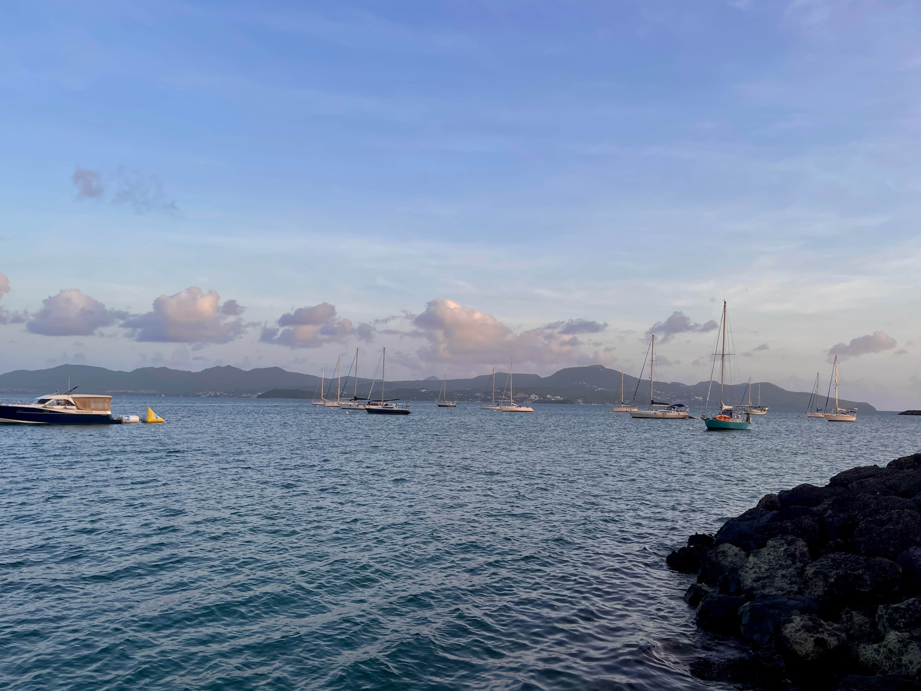 A calm harbor with several sailboats anchored, a rocky shoreline, and distant mountains under a partly cloudy sky.