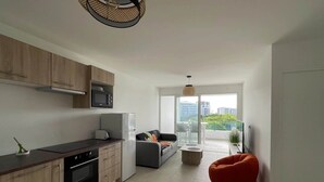 A modern kitchen with wooden cabinets, a microwave, and a refrigerator. There is a dining area with a sofa and a red bean bag chair. The room has a large window with a view of buildings and trees.