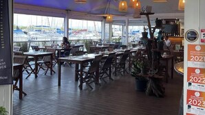 A restaurant with a view of boats through large windows, wooden tables and chairs, hanging pendant lights, and a sign indicating it is a bar and lounge.