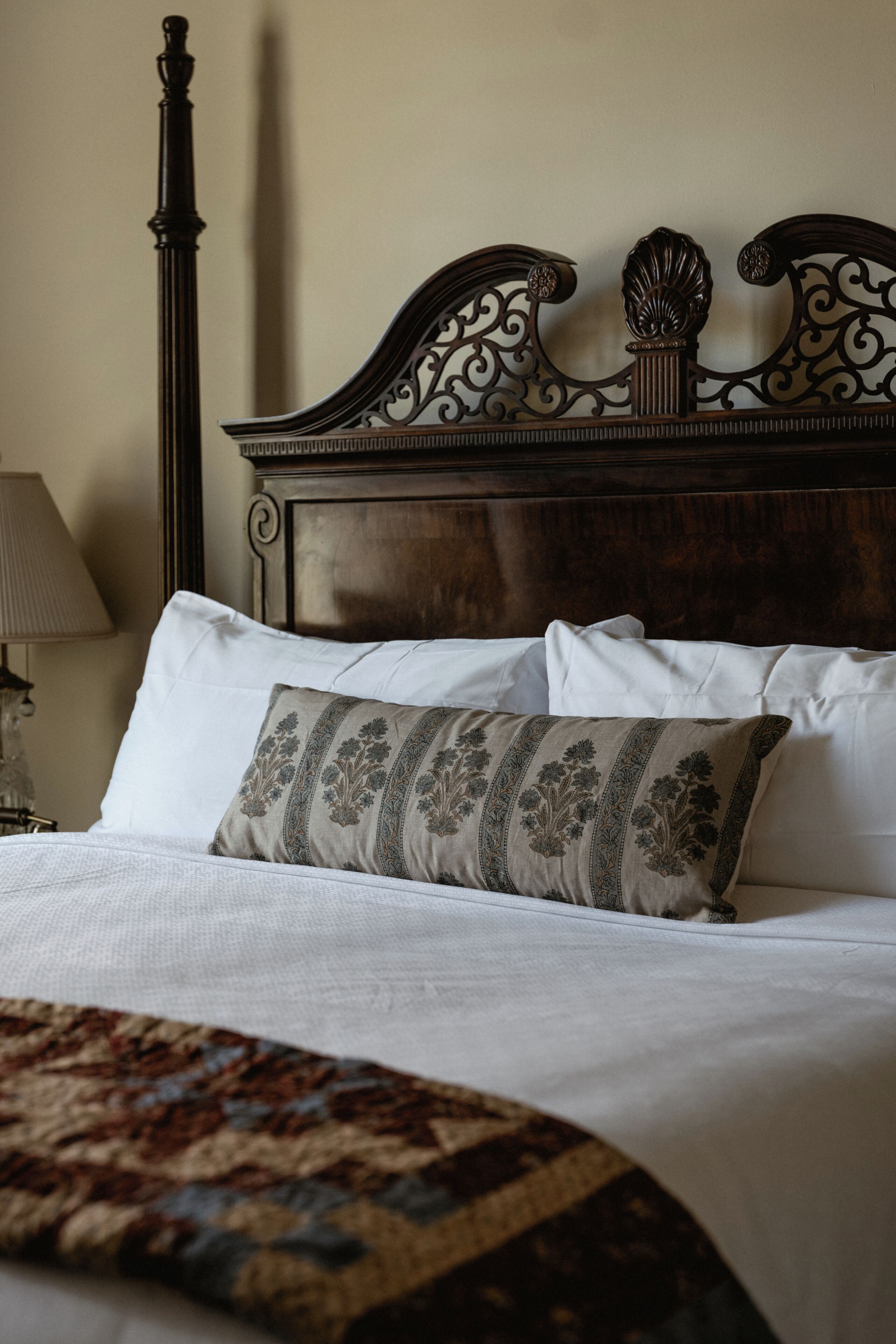 Una cama bien hecha con sábanas blancas y un cojín estampado, un cabecero de madera oscura con tallas intrincadas y una lámpara en una mesita de noche.