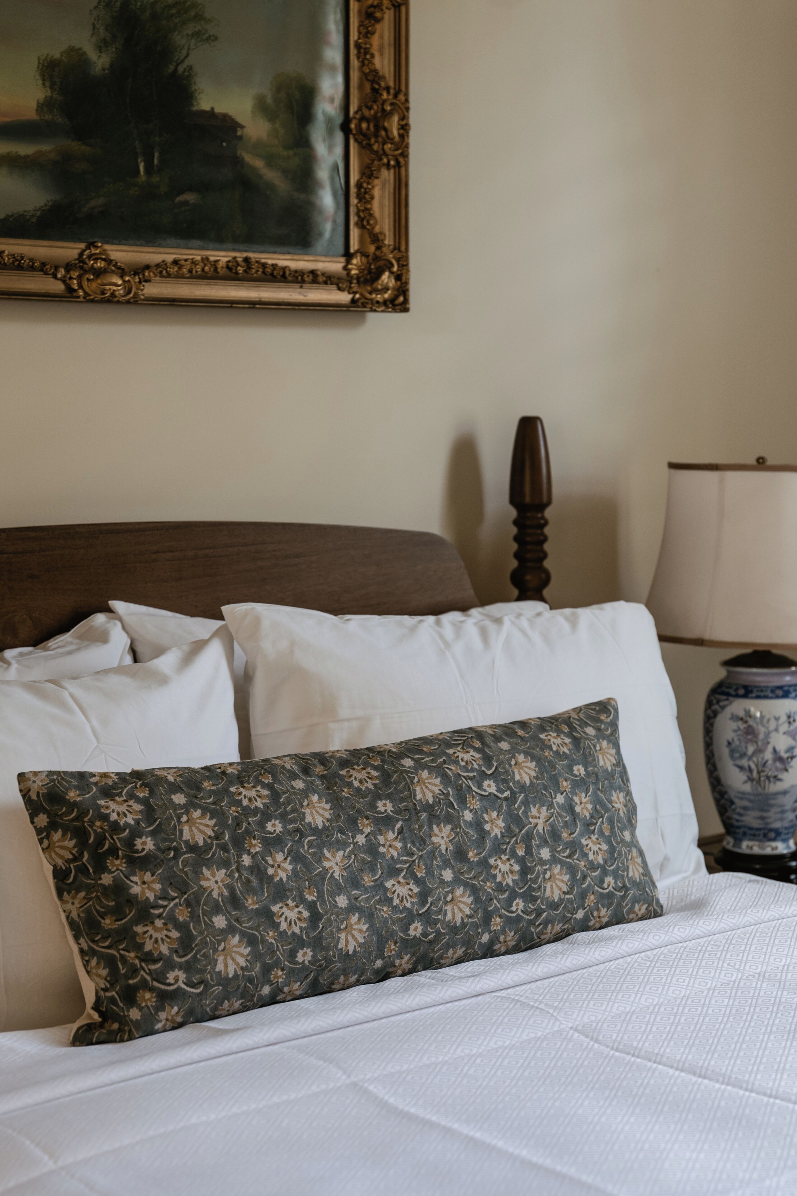 A bed with white and floral-patterned pillows, a wooden headboard, a vase with a floral design, and a painting on the wall.