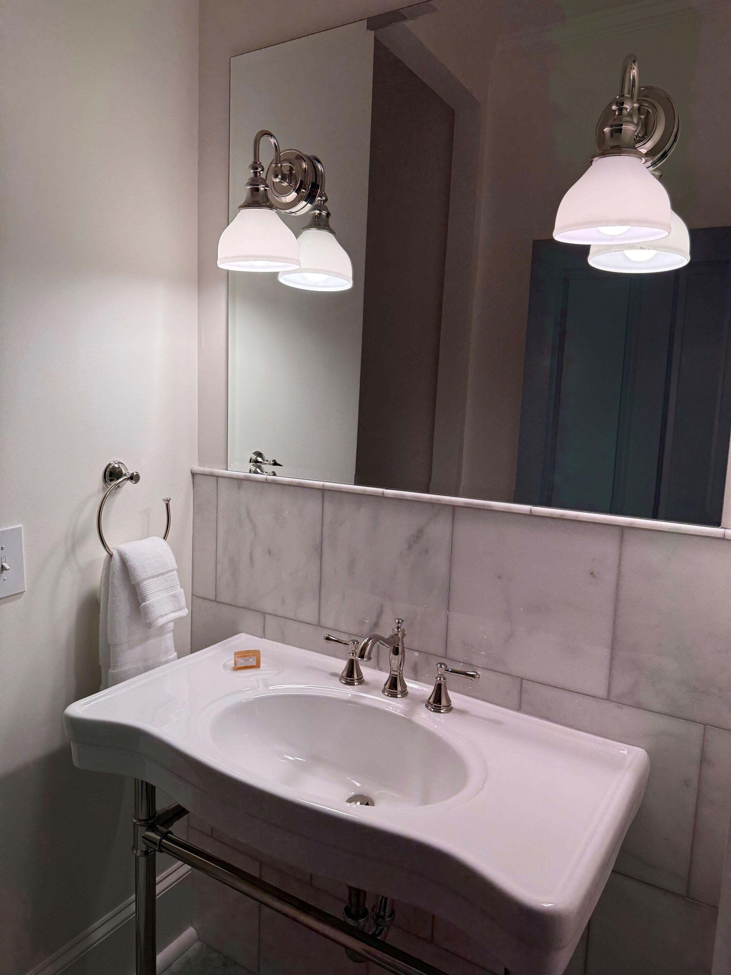 Un baño con un lavabo blanco, dos llaves, un perchero para toallas y un espejo con dos luces empotradas.