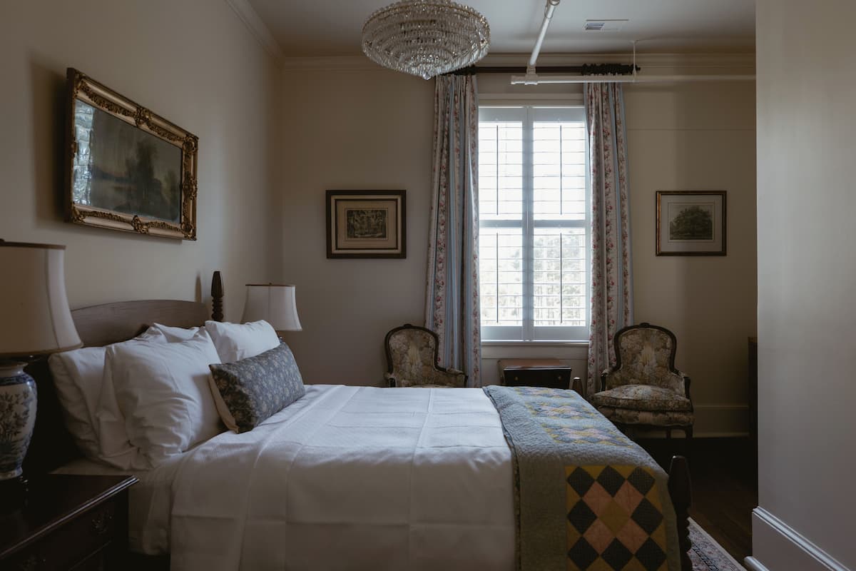 Una habitación de hotel con cama, dos sillas, una araña y una ventana con cortinas.