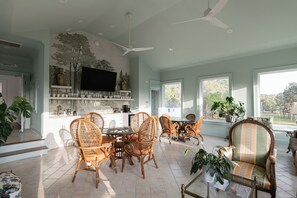 A spacious living area with a dining table surrounded by wicker chairs, a large armchair, and a television mounted on the wall.