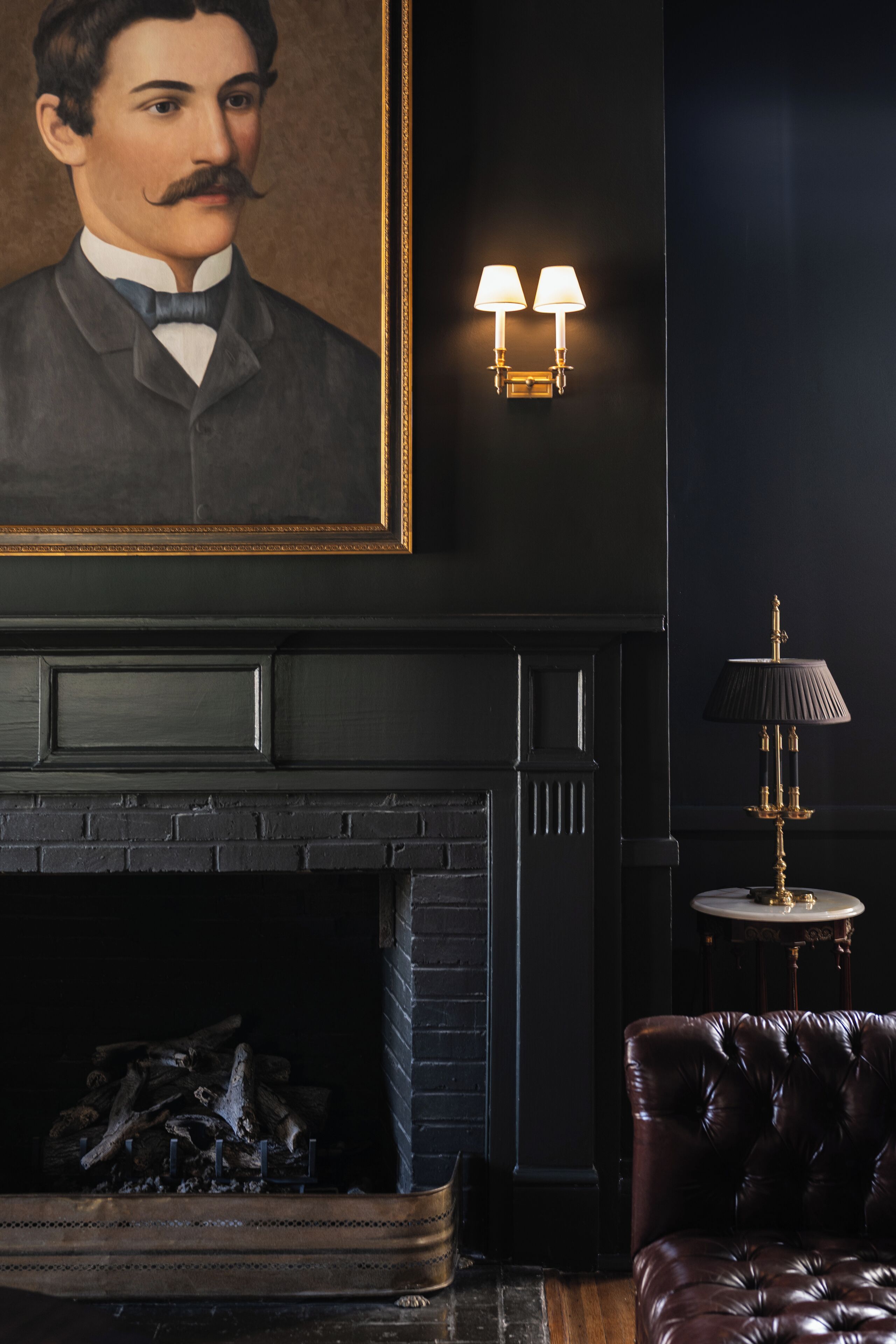 Una habitación de estilo oscuro con chimenea, un retrato enmarcado, lámparas de pared y un sillón de cuero.