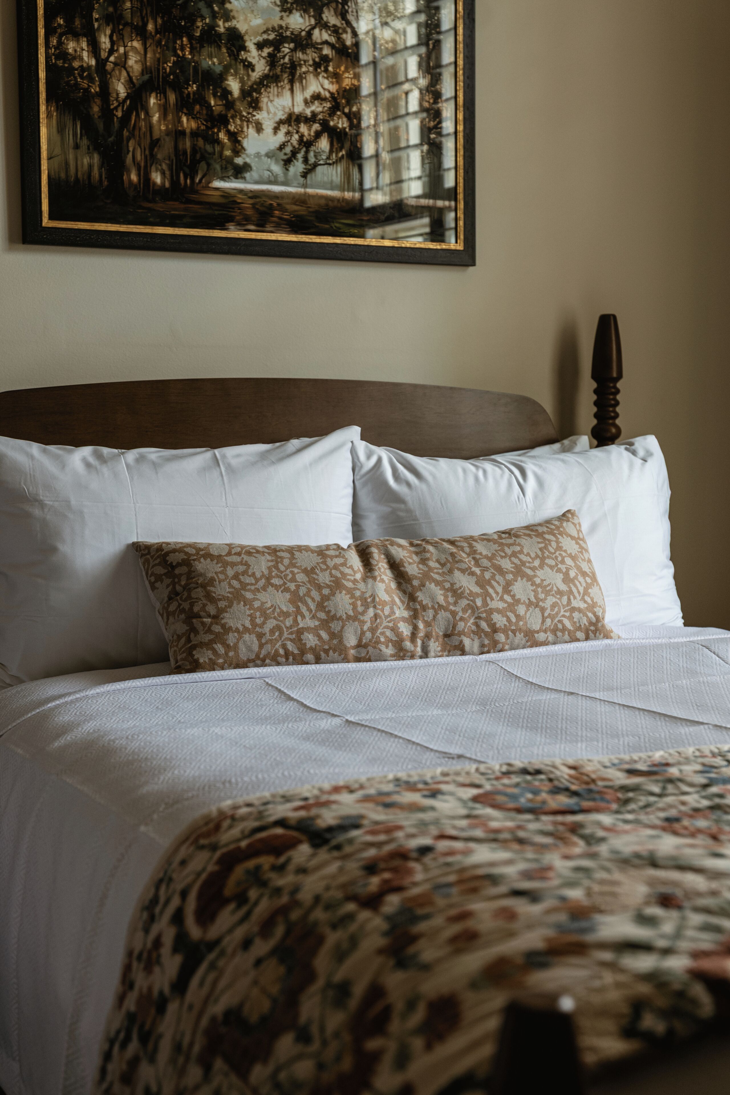 A neatly made bed with white linens and a patterned throw, a headboard, and a framed painting above.