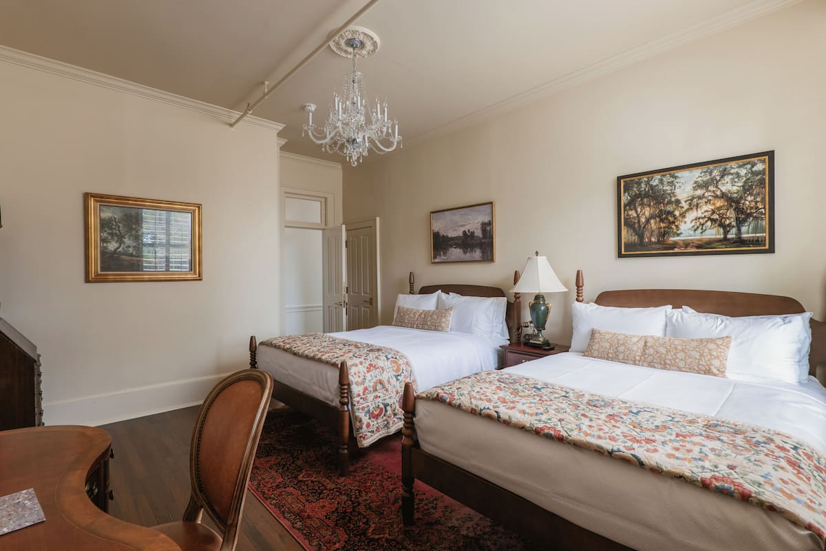 Habitación de hotel con dos camas, un candelabro, obras de arte enmarcadas y una silla de madera.