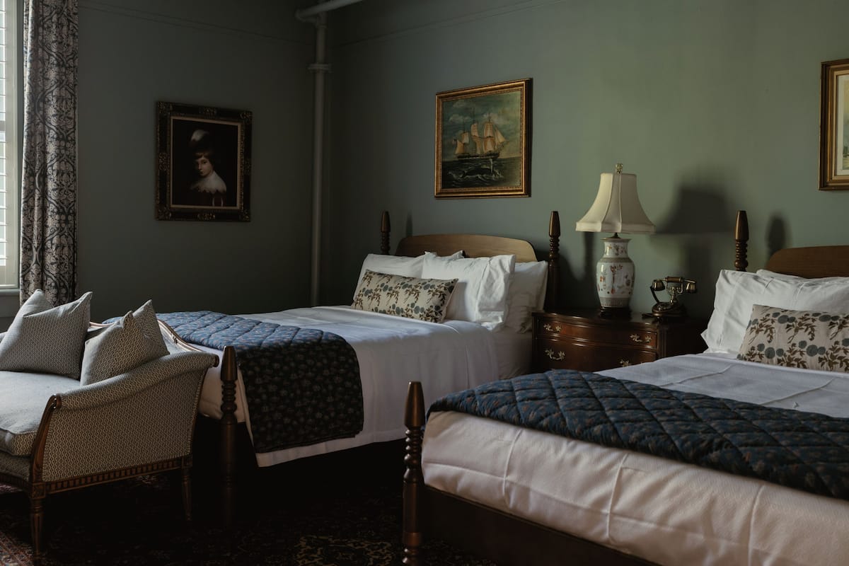 Una habitación con dos camas, una silla, una lámpara y dos cuadros en la pared.