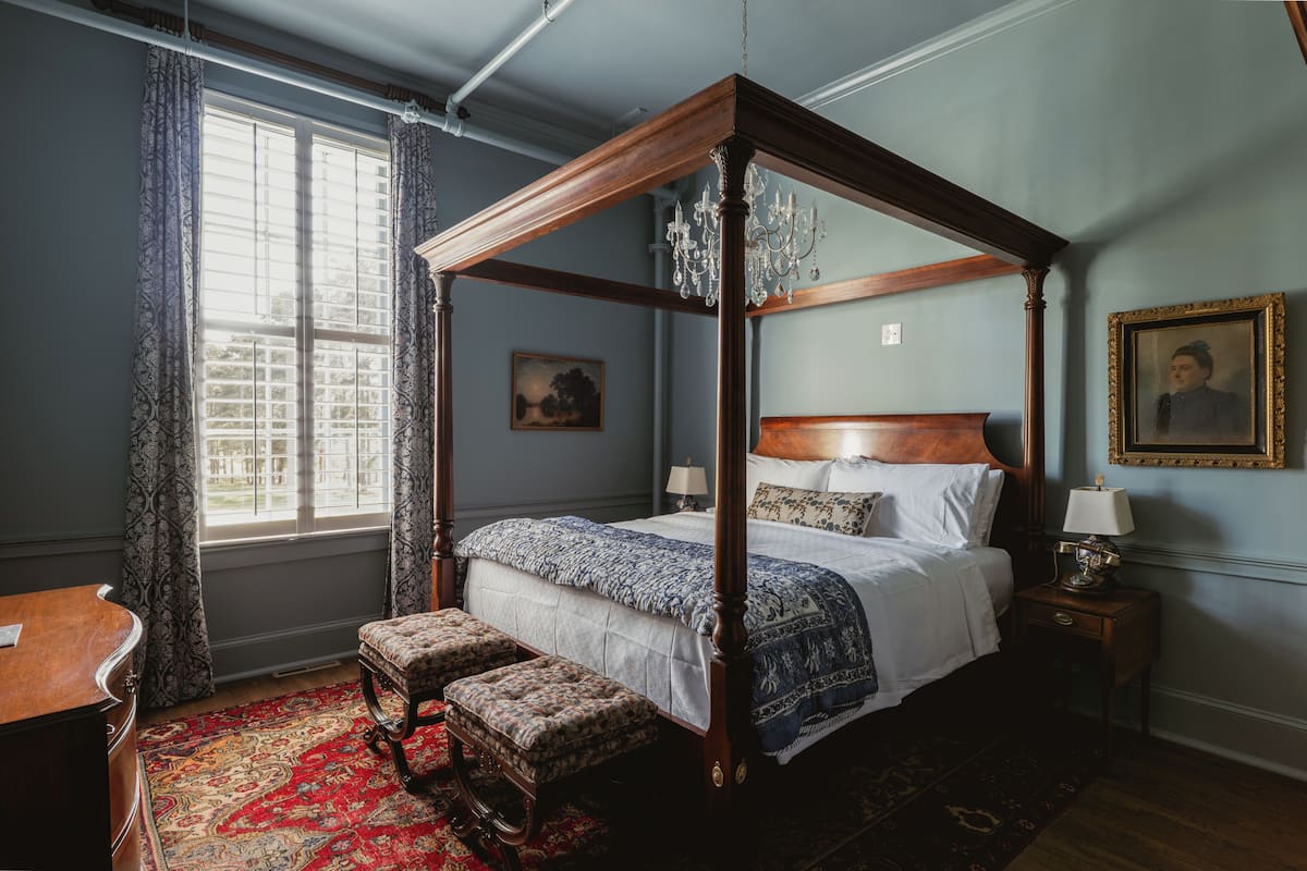 Una cama con dosel, un candelabro, una mesita de noche de madera con una lámpara, un banco y una mesa con una silla.