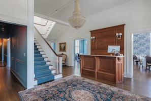 A spacious interior with a grand staircase, a wooden bar area, and a large chandelier.