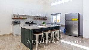 A modern kitchen with white cabinets, a central island with stools, a stainless steel refrigerator, and a window providing natural light.