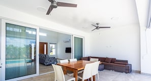 Amplia sala de estar con una mesa de comedor de madera, sillas blancas, un sofá marrón y un gran ventanal corredizo que da a un balcón.