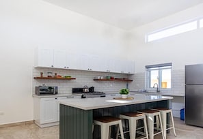Una cocina moderna con armarios blancos, una isla central con taburetes, un refrigerador de acero inoxidable y un ventanal que proporciona luz natural.
