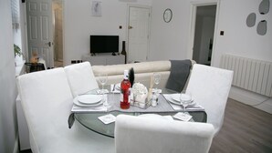 Un comedor con una mesa de vidrio, sillas blancas y un sofá blanco. Hay un televisor en la pared y una puerta que da a otra habitación.