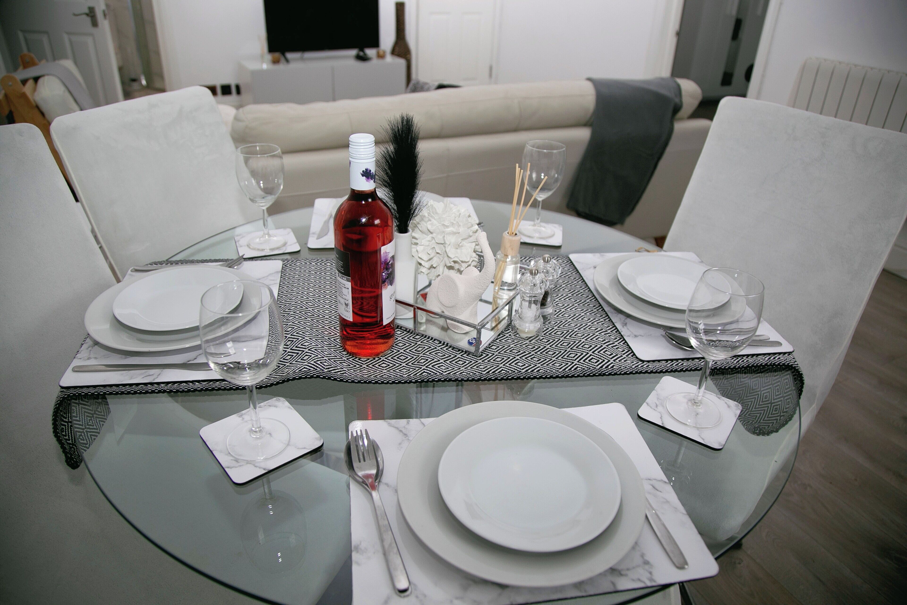 A dining table set for two with white plates, cutlery, and a bottle of red wine.