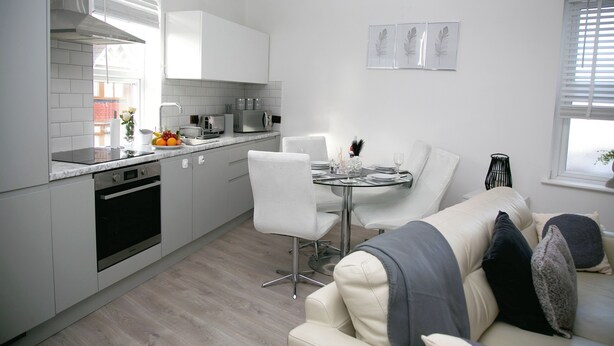 Una cocina moderna con armarios blancos, un comedor con una mesa redonda y sillas, y un sofá con cojines.