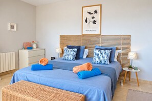 A bedroom with a bed, bedside tables, a radiator, a framed picture on the wall, and a wicker bench.