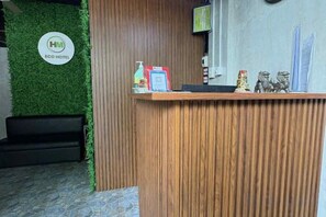 A reception desk with a wooden finish, a green wall with a logo, and a small statue on the counter. - HM Eco Hotel Naia (Pasay)