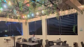 A dining area with tables and chairs, a person working at a desk, and large windows with blinds. - HM Eco Hotel Naia (Pasay)