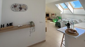 A spacious living area with a high ceiling, skylights, a wooden shelf with decorative items, and a view of a bedroom with a green sofa. - The Attic (Moya)