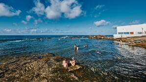 A coastal scene with people swimming and relaxing in the water, rocky shore, and a white building with a lifebuoy. - The Attic (Moya)