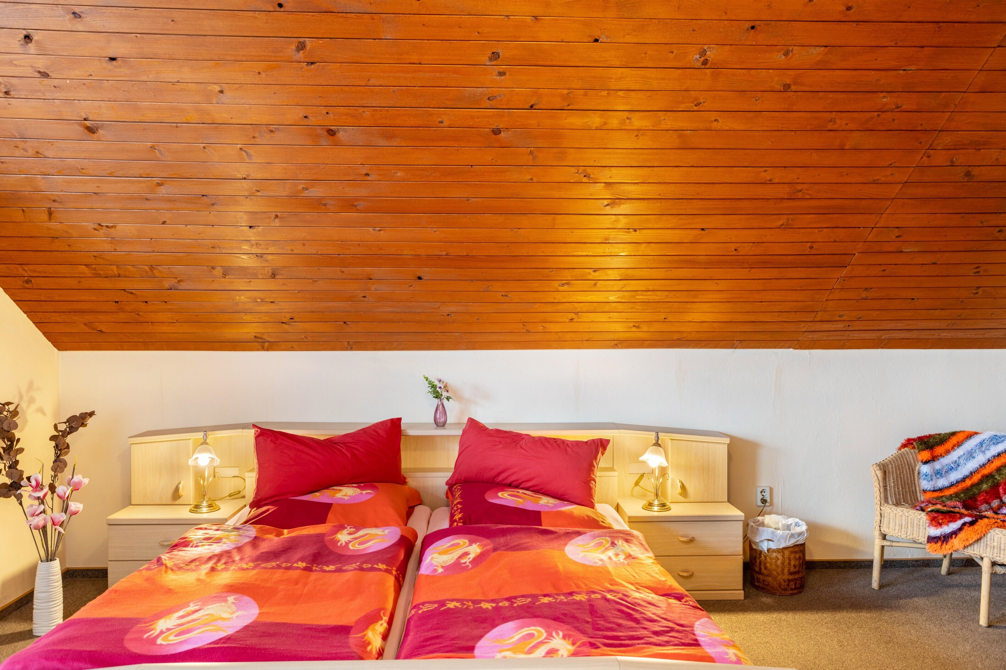 A room with a wooden ceiling, two beds with matching red bedding, bedside tables with lamps, a wicker chair with a blanket, and a vase with flowers.
