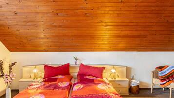 A room with a wooden ceiling, two beds with matching red bedding, bedside tables with lamps, a wicker chair with a blanket, and a vase with flowers.