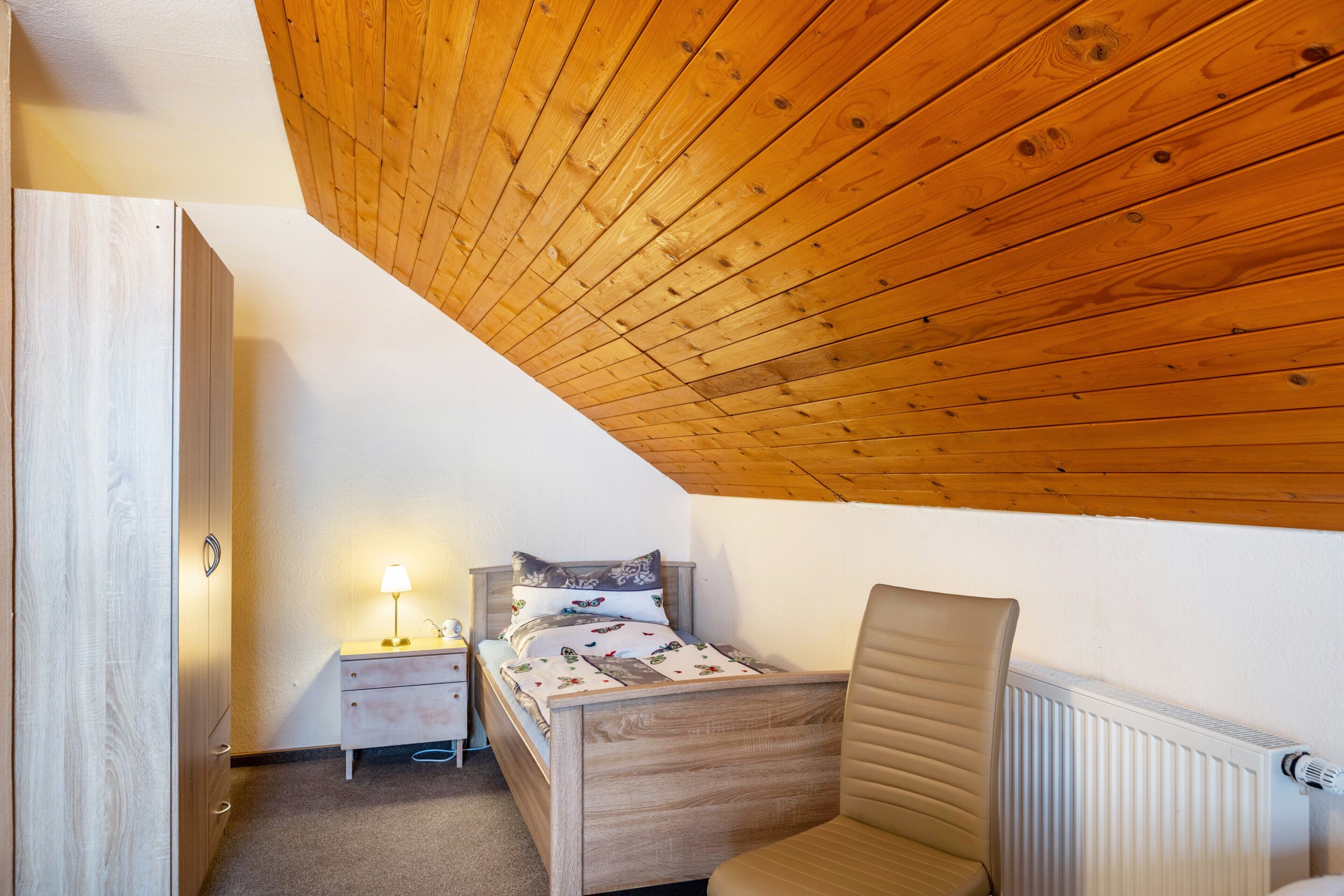 Una camera da letto con soffitto spiovente, un letto, un comodino, una sedia e un termosifone.