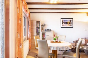 A dining area with a round table, chairs, a bookshelf, a framed picture, and a window with curtains. - Holiday Home 'Kaiser' with Mountain View, Private Terrace and Wi-Fi (Wasungen)