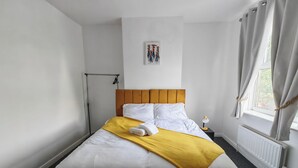 Un lit bien fait, avec une literie blanche et jaune, une tête de lit en bois, une table de chevet avec une lampe et une fenêtre avec des rideaux.