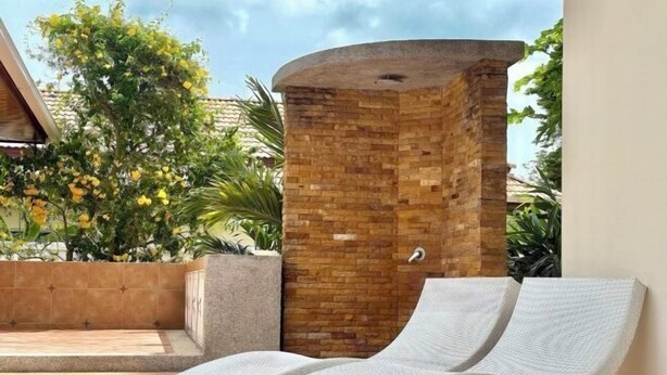 A sunroom with a brick wall, two lounge chairs, and a view of a tree with yellow flowers. - Chuan Pool Villa Rawai Phuket (Phuket)