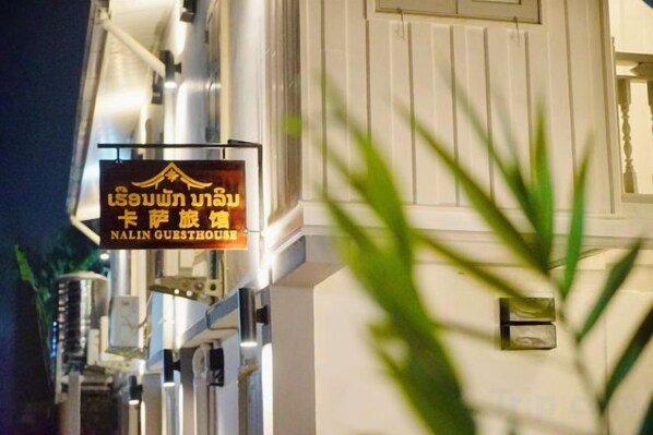 Schild für das Nalinn Guesthouse mit thailändischem Text, einer weißen Gebäudefassade und einer Pflanze im Vordergrund.