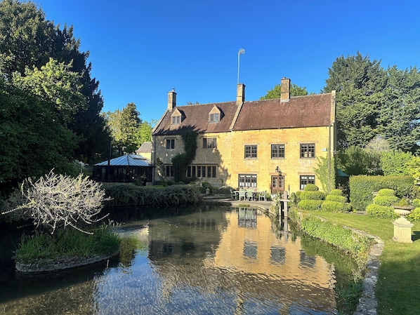 A historic stone building with a reflective pond, surrounded by lush greenery and a clear blue sky.